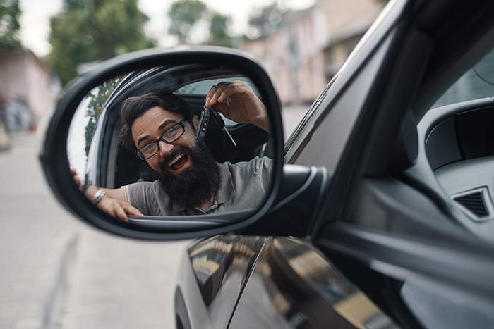 Car dealership, the happiest client. Charismatic man holding car keys smiling, with teeth, while sitting in the car looking to himself in the side mirror reflection.